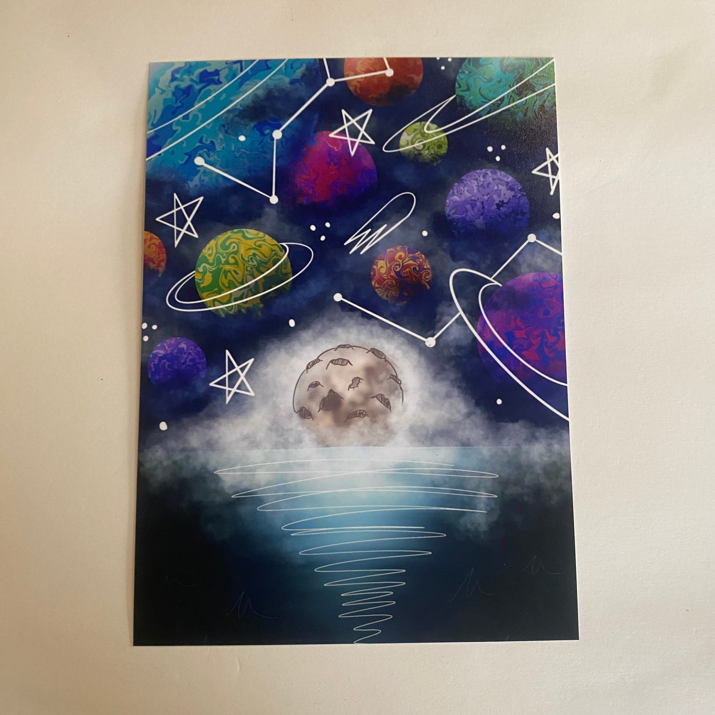 A dark blue sky is filled with colorful planets with swirly designs and encases in dark blue and white clouds. A large moon rests on the dark teal-blue waters. The piece is covered with simple line-art illustrations such as classic stars, constellations, comets, and rings, making the piece appear to have been drawn on top of with a marker.